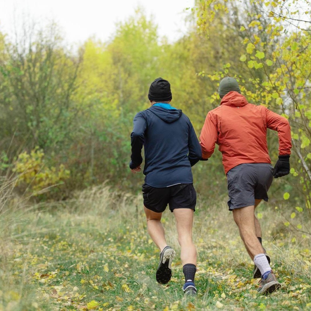 wei Männer joggen durch einen herbstlichen Wald. Beide tragen die AchilloTrain Bandage von Bauerfeind, die für Stabilität und Unterstützung sorgt.