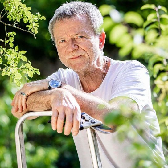 Dieter verschneidet Gewächse in seinem Garten. Sein Garten ist seine große Leidenschaft. Er hält eine Gartenschere in seiner rechten Hand und lächelt in die Kamera.