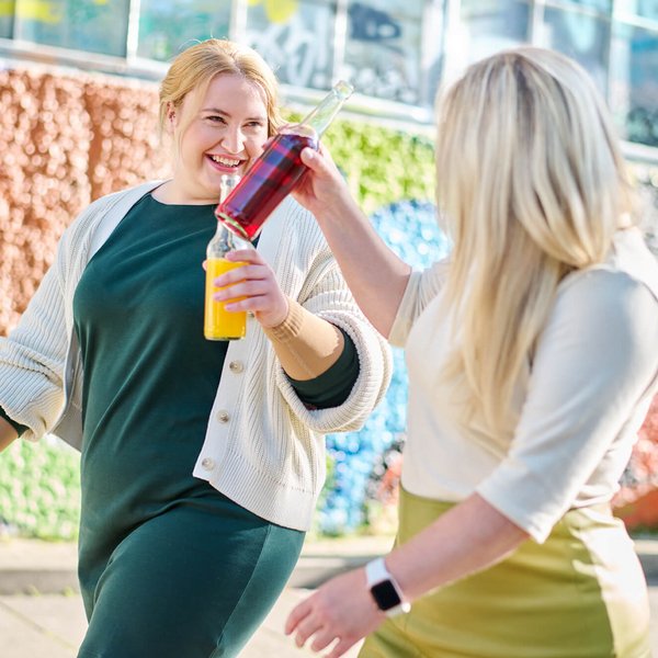 Zwei Frauen stoßen mit farbenfrohen Getränkeflaschen an, während sie lächelnd durch eine urbane Umgebung spazieren. Im Hintergrund ist eine bunte Wand mit grafischen Mustern zu sehen, die eine lebendige Atmosphäre schafft.
