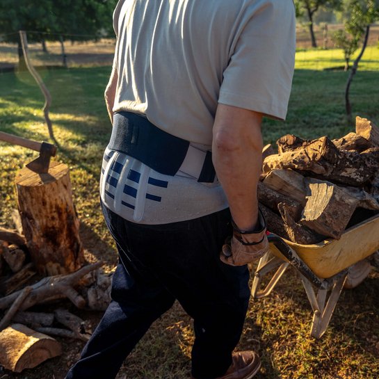 Ein Mann mit Rückenbandage von hinten, beim Bewegen einer gelben Schubkarre voller Holzstücke im Garten. Im Hintergrund steht ein Baumstumpf mit einer Axt, umgeben von Holzscheiten.