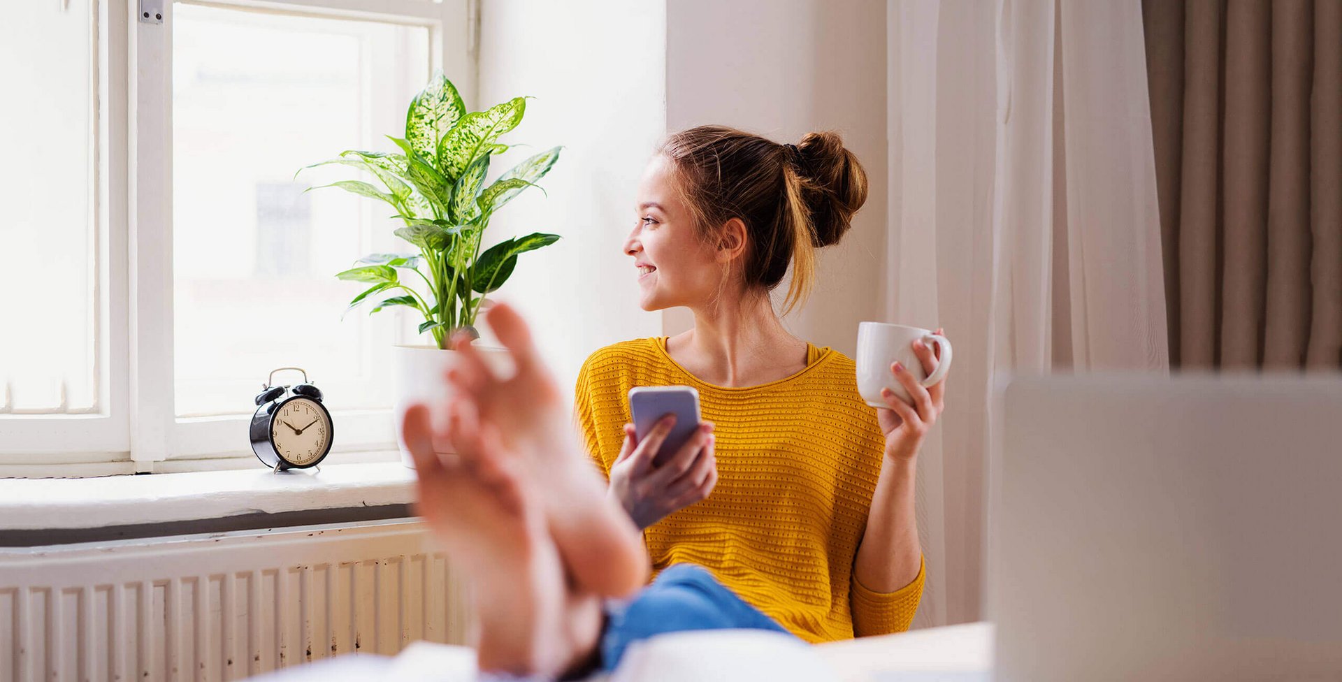 Auf dem Bild sitzt eine junge Frau auf einem Stuhl und hat ihre Füße auf den Tisch vor ihr abgelegt. In der linken Hand hält sie eine Tasse; in der rechten Hand ein Smartphone. Regelmäßiges Training der Fußmuskulatur kann Schmerzen vorbeugen.