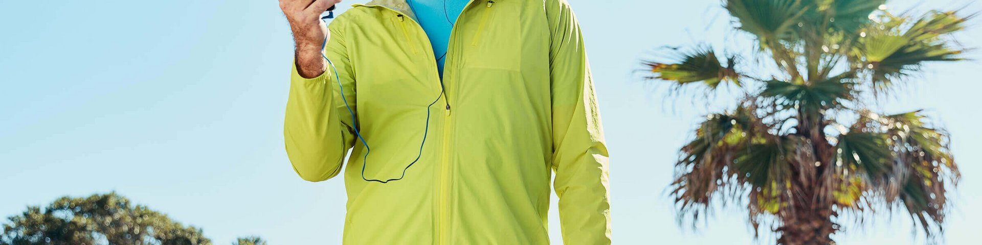 Ein älterer Mann in sportlicher Kleidung mit einem gelben Sportjackett und einer schwarzen Kappe steht draußen unter klarem Himmel. Er hält ein Smartphone in der Hand und trägt blaue In-Ear-Kopfhörer. 