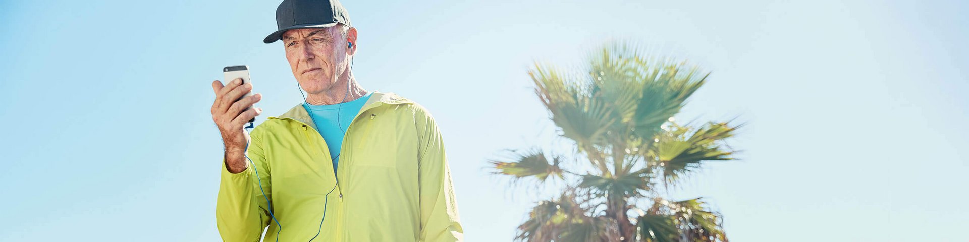 Ein älterer Mann in sportlicher Kleidung mit einem gelben Sportjackett und einer schwarzen Kappe steht draußen unter klarem Himmel. Er hält ein Smartphone in der Hand und trägt blaue In-Ear-Kopfhörer. 