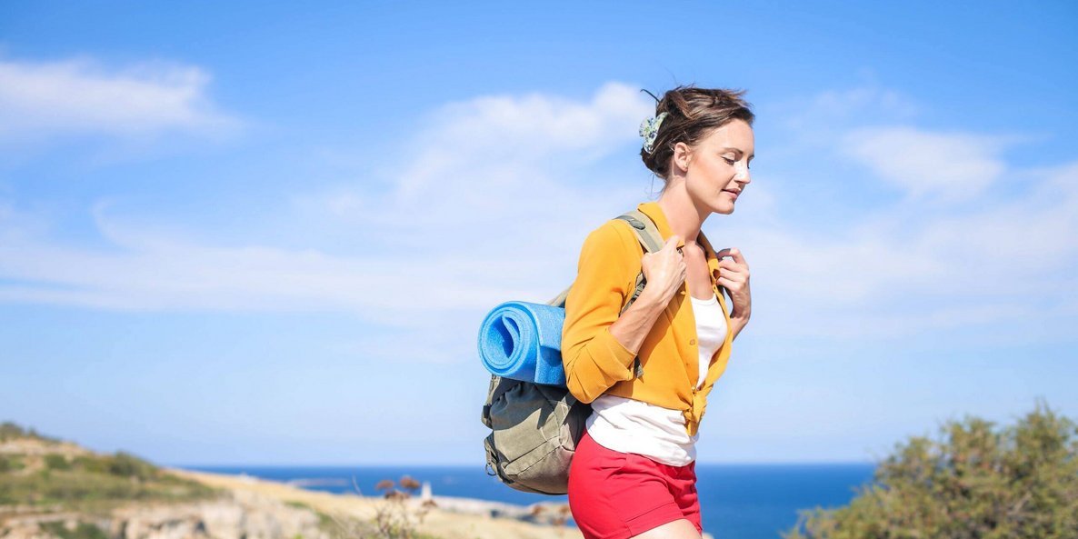 Das Bild zeigt eine junge Frau mit einem Wanderrucksack bei Sonnenschein am Meer. In den warmen Sommermonaten fällt es uns meist leichter, aktiv zu werden und Bewegungsmangel einzudämmen.