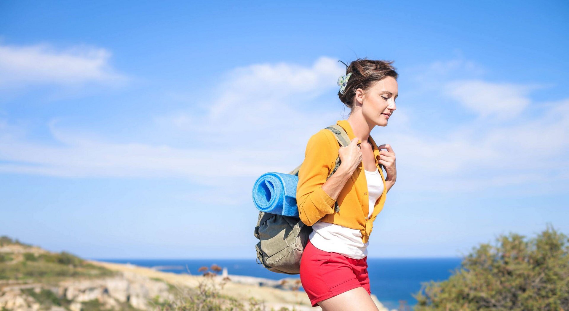 Das Bild zeigt eine junge Frau mit einem Wanderrucksack bei Sonnenschein am Meer. In den warmen Sommermonaten fällt es uns meist leichter, aktiv zu werden und Bewegungsmangel einzudämmen.