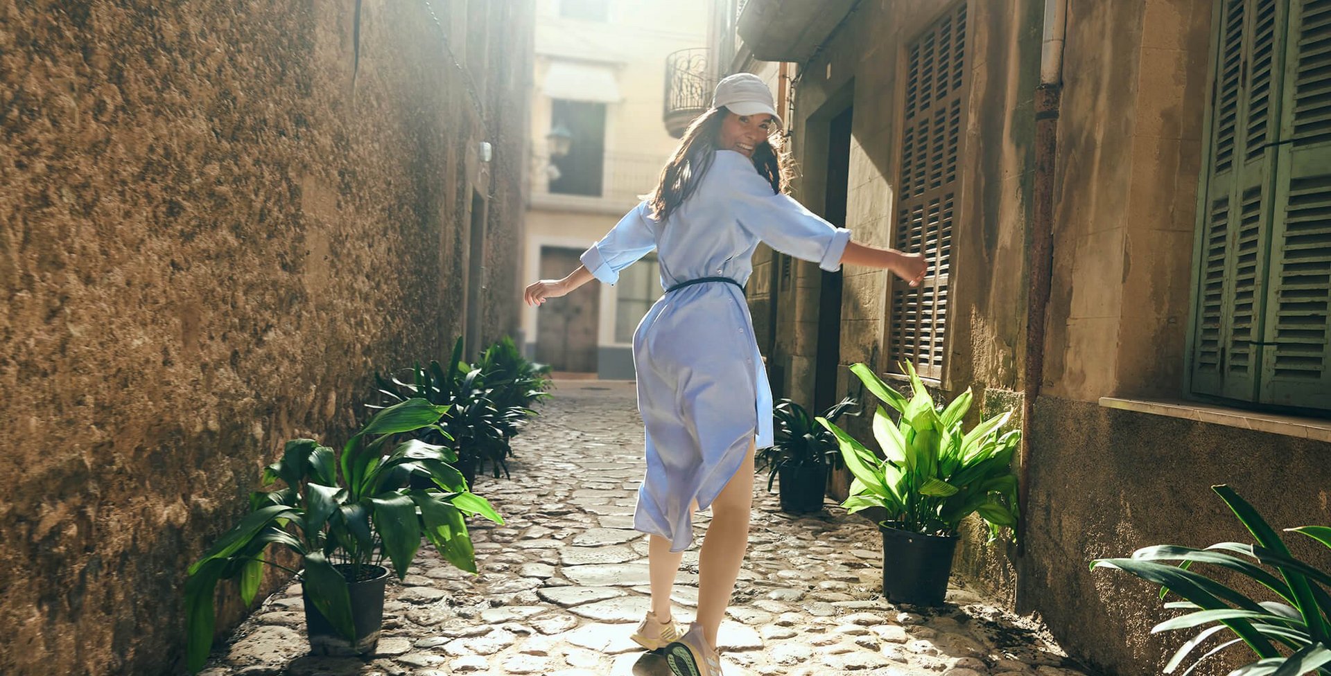 Eine junge Frau in einem hellblauen Kleid und Sneakers läuft fröhlich durch eine enge, mit Kopfsteinpflaster versehene Gasse. Die Sonne scheint durch die alten Gebäude und erzeugt ein warmes Licht. 