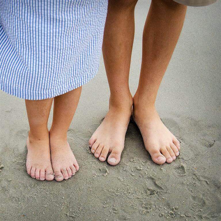 Bildausschnitt mit zwei Kinderbeinen, die barfuß auf sandigem Boden stehen. Regelmäßiges Barfußlaufen kann zur Fußgesundheit beitragen.