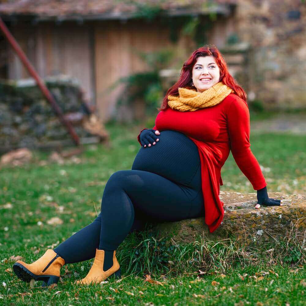 Das Bild zeigt Lipödem-Patientin Kathi Schanz während ihrer Schwangerschaft, die lächelnd auf einem Stein im Garten sitzt. Zur Unterstützung trägt sie ihre VenoTrain curaflow Versorgungen.