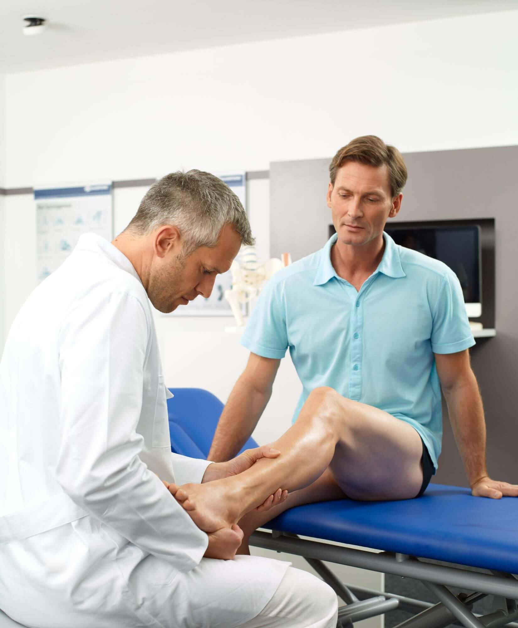 Auf dem Bild untersucht ein Arzt das Sprunggelenk eines Patienten in seinem Behandlungszimmer. Bei einer Sprunggelenksdistorsion sollte das betroffene Fußgelenk umgehend ruhig gestellt, gekühlt und hochgelagert werden.